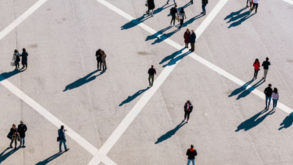 Aerial photo of scattered pedestrians crossing a large plaza, long diagonal shadows suggest movement and urban scale.